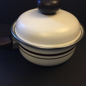 May include: A white enamel saucepan with a lid and a dark brown handle. The pot has two dark brown stripes around the body and a dark brown knob on the lid. The pot is made of metal and is designed for cooking.