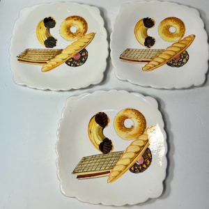 May include: Three square, white ceramic plates with scalloped edges. Each plate features a hand-painted design of a bagel, croissant, baguette, and a pastry. The food illustrations are in shades of brown, yellow, and pink.