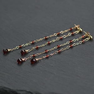 May include: Gold-toned dangle earrings featuring a series of small, round, deep red beads strung along delicate chains. The earrings have a simple, elegant design with a single larger bead at the end of each chain. The earrings are displayed against a dark background.