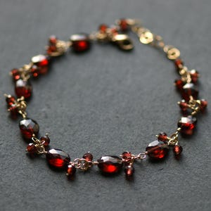 May include: A gold bracelet with deep red gemstones. The bracelet features a delicate chain with clusters of smaller stones and larger, oval-shaped gems. The clasp is visible at the end of the chain. The gemstones are a rich, dark red colour.
