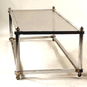 May include: A rectangular coffee table featuring a clear glass top surface supported by a polished chrome metal frame. The structure includes four vertical cylindrical legs topped with spherical finials and a matching base with ball feet.