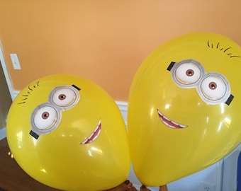 May include: Two yellow balloons with Minion faces printed on them. The Minions have black eyebrows, black goggles, and white eyes with brown pupils. One Minion is smiling with a white mouth and teeth. The other Minion has a white mouth with a black line for a smile.