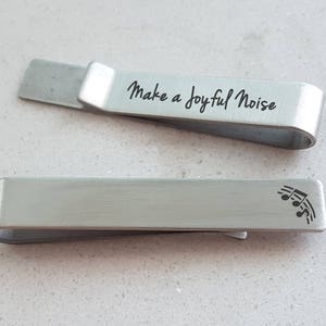 Könnte beinhalten: Zwei silberne Krawattenklammern aus Metall. Eine Krawattenklammer hat den Text "Make a Joyful Noise" eingraviert. Die andere Krawattenklammer hat ein Musiknotensymbol eingraviert.