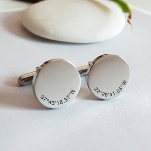 May include: A pair of silver cufflinks with engraved coordinates. The left cufflink reads "37°43'18.37"N" and the right cufflink reads "22°28'41.05"W".