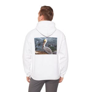 May include: White hooded sweatshirt with a rectangular image of a pelican on the back. The pelican has a long yellow beak and is perched near the ocean. The sweatshirt is a solid white color.