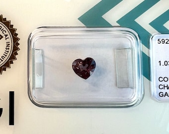 May include: A heart-shaped gemstone in a clear plastic case. The gemstone is a deep reddish-purple color.