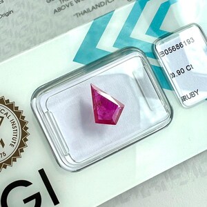 Könnte beinhalten: Ein facettierter, schildförmiger Rubin mit leuchtend pink-rotem Farbton, sicher untergebracht in einer transparenten Kunststoff-Präsentationsbox. Die Box liegt auf einem weißen Zertifikat mit dem Text B05686193, 0.90 Ct und RUBY.