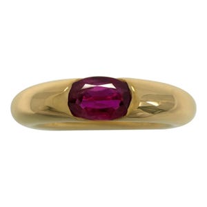 May include: A gold ring featuring a vibrant, oval-cut ruby gemstone. The ring has a smooth, polished band and a classic, elegant design. The ruby is a deep, rich red color, set in a simple, yet striking setting.