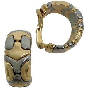 May include: A pair of gold and silver-tone hoop earrings. The earrings feature a geometric design with interlocking segments of gold and silver metal. The earrings have a hinged clasp for secure wear.