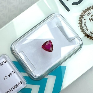 Peut inclure: Un rubis rouge vif taille trillion présenté dans un boîtier en plastique transparent. Le boîtier est posé sur une carte blanche portant l'inscription 0.44 Ct et 80566760, ainsi qu'un sceau circulaire marqué International Institute 1975.