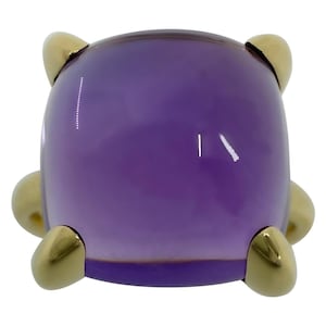 Pode incluir: Um anel com uma grande pedra preciosa de ametista roxa lisa, lapidada em formato de cabochão arredondado. A pedra é mantida firmemente no lugar por quatro garras de metal dourado polido que se estendem da base, emoldurando a pedra roxa.