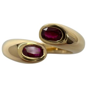 May include: A gold-toned ring featuring two oval-shaped, deep red gemstones. The ring's design is a continuous, flowing form, with each gemstone set in a polished gold bezel. The ring is set against a white background.