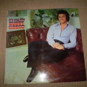 May include: A vintage vinyl record album cover featuring a man in a light blue shirt and dark pants sitting on a brown leather couch. The album title is "it's my life BUDDY GRECO".
