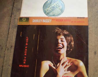 Op de afbeelding: Een vintage vinyl platenhoes met Shirley Bassey met Nelson Riddle en zijn orkest. De hoes is rood, zwart en wit met een foto van Shirley Bassey die zingt. De titel van het album is "Let's Face the Music".