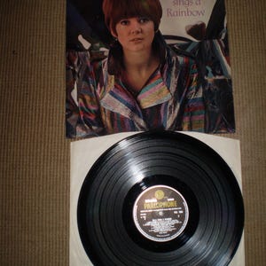 Könnte beinhalten: Ein Vintage-Vinyl-Schallplattencover mit Cilla Black, einer britischen Sängerin. Das Cover ist eine Nahaufnahme von Cilla, die einen farbenfrohen gestreiften Mantel trägt und direkt in die Kamera schaut. Der Albumtitel lautet "Cilla sings a Rainbow" und ist in weißer Schrift auf schwarzem Hintergrund gedruckt.