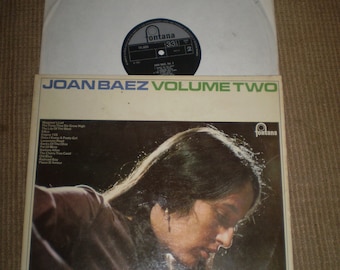 May include: A vinyl record album cover featuring Joan Baez. The cover is a close-up of Joan Baez's face, with her hair covering most of her face. The album title "JOAN BAEZ VOLUME TWO" is printed in large, bold letters at the top of the cover. The album cover is a light brown color with a white border.