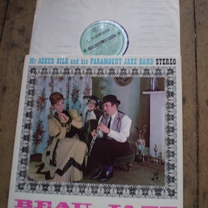 Peut inclure: Une pochette d'album de disque vinyle vintage mettant en scène Mr. Acker Bilk et son Paramount Jazz Band. La pochette présente une photo du groupe dans un salon avec une femme en robe et un homme jouant de la clarinette. Le titre de l'album est "Beau Jazz" en lettres roses.