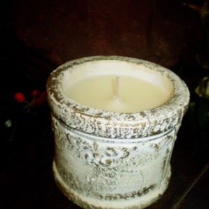 May include: A cream-coloured candle in a distressed, off-white ceramic container with gold accents. The candle has a single wick. The container has a textured, aged appearance and is set against a dark, blurred background.
