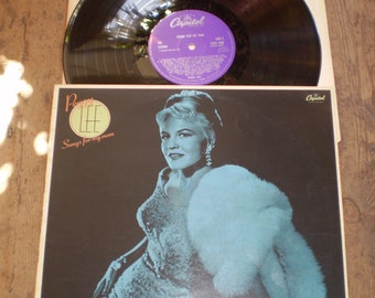 Op de afbeelding: Een vintage vinyl platenhoes met een vrouw in een witte bontjas en een paillettenjurk. Het album is getiteld "Peggy Lee Sings for my man" en heeft het Capitol Records-logo.
