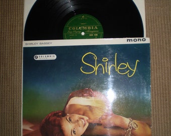 Op de afbeelding: Een vintage vinyl platenhoes met een foto van Shirley Bassey, een zangeres, liggend op een lichtgekleurd oppervlak. De albumhoes is blauw met de titel "Shirley" in gele cursieve letters. Het platenlabel is Columbia Records.