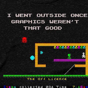 Op de afbeelding: Donkergrijs t-shirt met de tekst "I WENT OUTSIDE ONCE GRAPHICS WEREN'T THAT GOOD." Het shirt heeft een retro videogame-afbeelding met een pixelontwerp in geel, groen, blauw, rood en paars.