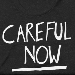 Pode incluir: T-shirt cinzenta escura com as palavras "CAREFUL NOW" em letras maiúsculas brancas grandes. Uma linha branca sublinha a palavra "NOW". A t-shirt está cuidadosamente dobrada.