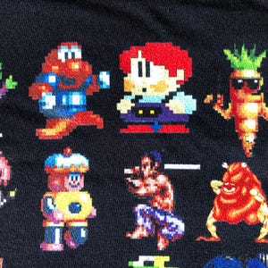 May include: Black fabric featuring a pixelated design of various video game characters. The characters are in bright colors, including red, green, blue, and yellow. The design is reminiscent of classic 8-bit video games.