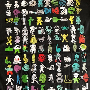 Puede incluir: Camiseta negra con un diseño pixelado de varios personajes y objetos coloridos. El diseño incluye una variedad de figuras, como una persona, un perro, un oso, un coche, una nave espacial, una araña, un pájaro, un pez, un hongo, una flor, un árbol, una casa, una estrella, un corazón, un círculo, un cuadrado, un triángulo, una línea, un punto y una letra.