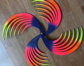 May include: A kinetic sculpture featuring four curved, fan-like blades attached to a central pivot point. Each blade transitions from a dark blue base to a vibrant gradient of neon orange and yellow at the tips. The structure is symmetrical.