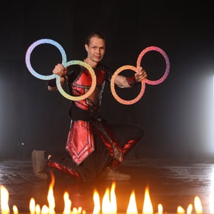 May include: A man in a red and black costume is performing with three iridescent hoops. He is kneeling on a stage with flames in the foreground.