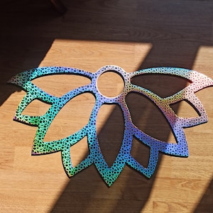 Peut inclure: Une décoration murale plate en forme de fleur de lotus stylisée avec un centre circulaire. La surface présente une finition holographique iridescente avec un motif répétitif de petits cercles superposés qui reflètent des couleurs arc-en-ciel.