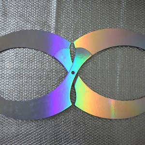 Puede incluir: Un objeto plateado en forma de infinito con secciones iridiscentes de color arcoíris. El objeto parece ser de metal y tiene bordes curvos. Está sobre un fondo plateado texturizado.