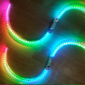 Pode incluir: Quatro tiras de luz LED curvas dispostas em um padrão em forma de oito. As luzes transitam por um espectro de cores, incluindo rosa, verde e amarelo. As luzes estão em uma superfície de madeira.