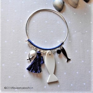 Peut inclure: Un bracelet jonc argenté avec un cordon bleu et des breloques. Les breloques comprennent un poisson blanc, un pompon bleu, une perle blanche et une perle noire. Le bracelet est sur un fond blanc.