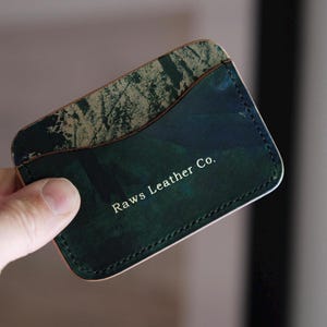 Op de afbeelding: Een leren kaarthouder met een groen en bruin gemarmerd ontwerp. De kaarthouder heeft de tekst "Raws Leather Co." op de voorkant. De randen zijn gestikt met een contrasterende draad.