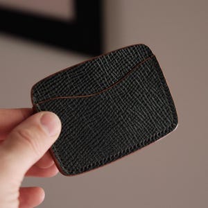 以下が含まれることがあります： 黒い革製のカードケース。表面は質感があり、縁は茶色。カードケースには前面にポケットがあり、人の手で持たれています。