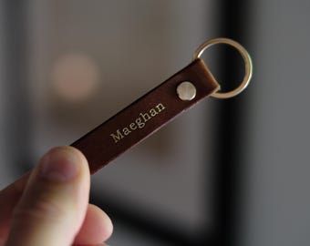 May include: A brown leather keychain with a gold-colored key ring and a silver rivet. The name "Maeghan" is embossed in gold lettering on the leather strap. The keychain is held in a person's hand.