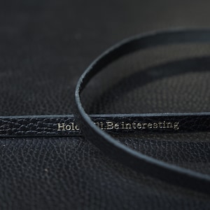 Peut inclure: Gros plan sur un bracelet en cuir noir avec l'inscription gravée "Hold 1. Be interesting". Le bracelet est circulaire et repose sur une surface noire texturée. L'image met en valeur le design épuré du bracelet et le message gravé.