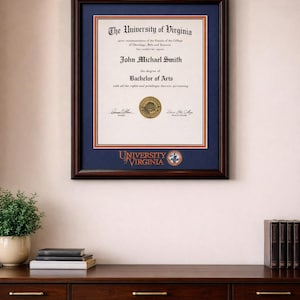 May include: A framed diploma from The University of Virginia, featuring the name John Michael Smith and the degree of Bachelor of Arts. The diploma is set against a navy blue mat within a dark wood frame. A brass lamp is mounted above the frame.