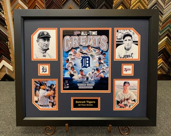 May include: Framed artwork featuring Detroit Tigers baseball legends. The display includes portraits, team logos, and the text "ALL-TIME GREATS." The frame is black, with a dark blue matting and orange accents. The artwork is mounted on a metal stand.