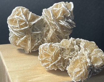 May include: Two beige rose quartz crystals, also known as desert roses, on a light brown wooden surface. The crystals are clustered together and have a delicate, layered structure.