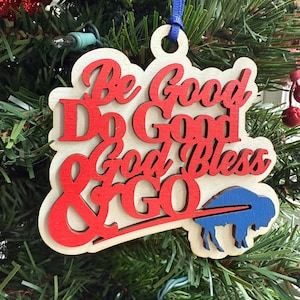 May include: Wooden Christmas ornament with the words "Be Good Do Good God Bless & GO" in red script. A blue buffalo silhouette is on the bottom right. The ornament hangs from a blue ribbon and is displayed on a Christmas tree.