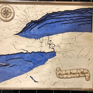 Pu&ograve; includere: Una mappa in legno della regione di Buffalo Niagara con elementi d'acqua blu e una rosa dei venti in alto a sinistra. La mappa &egrave; incorniciata in una cornice di legno scuro.