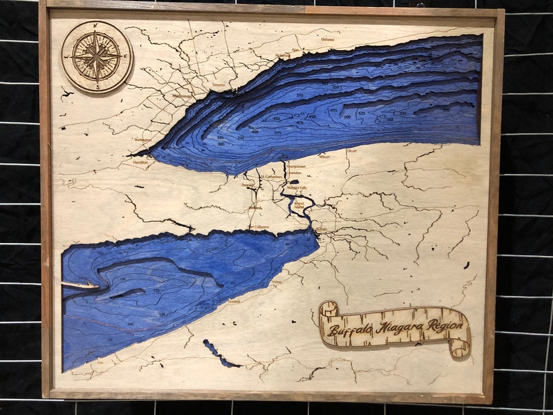 Pu&ograve; includere: Una mappa in legno della regione di Buffalo Niagara con elementi d'acqua blu e una rosa dei venti in alto a sinistra. La mappa &egrave; incorniciata in una cornice di legno scuro.