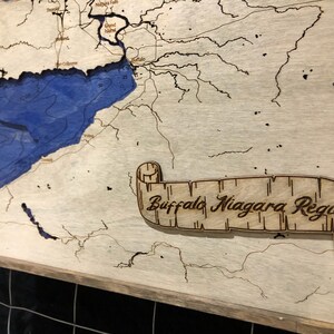 Pu&ograve; includere: Una mappa in legno della regione di Buffalo Niagara con un lago e fiumi blu. La mappa &egrave; incorniciata e ha un rotolo con il testo "Buffalo Niagara Region".