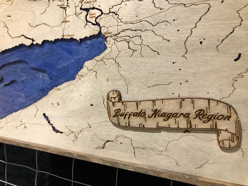 Pu&ograve; includere: Una mappa in legno della regione di Buffalo Niagara con un lago e fiumi blu. La mappa &egrave; incorniciata e ha un rotolo con il testo "Buffalo Niagara Region".