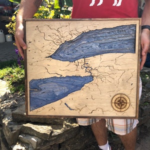 Pu&ograve; includere: Una mappa in legno del Lago Erie con linee di profondit&agrave; blu e una rosa dei venti. La mappa &egrave; incorniciata in una cornice di legno scuro ed &egrave; tenuta da una persona.