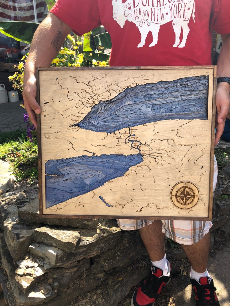 Pu&ograve; includere: Una mappa in legno del Lago Erie con linee di profondit&agrave; blu e una rosa dei venti. La mappa &egrave; incorniciata in una cornice di legno scuro ed &egrave; tenuta da una persona.
