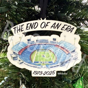 May include: A Christmas tree ornament featuring a watercolor illustration of a football stadium. The ornament reads "THE END OF AN ERA" above the stadium and "1973-2025" below. The stadium has blue, red, and green colors.