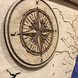Pu&ograve; includere: Una bussola in legno con un design dettagliato, che presenta le lettere N, S, E e W. La bussola &egrave; posizionata su uno sfondo in legno chiaro con una mappa blu e bianca.